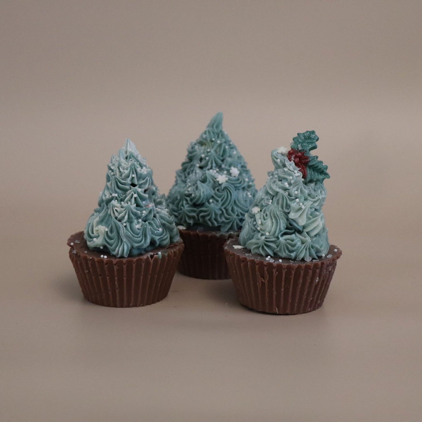 Three chocolate cupcake soaps with dark green frosting shaped like Christmas trees on a beige background