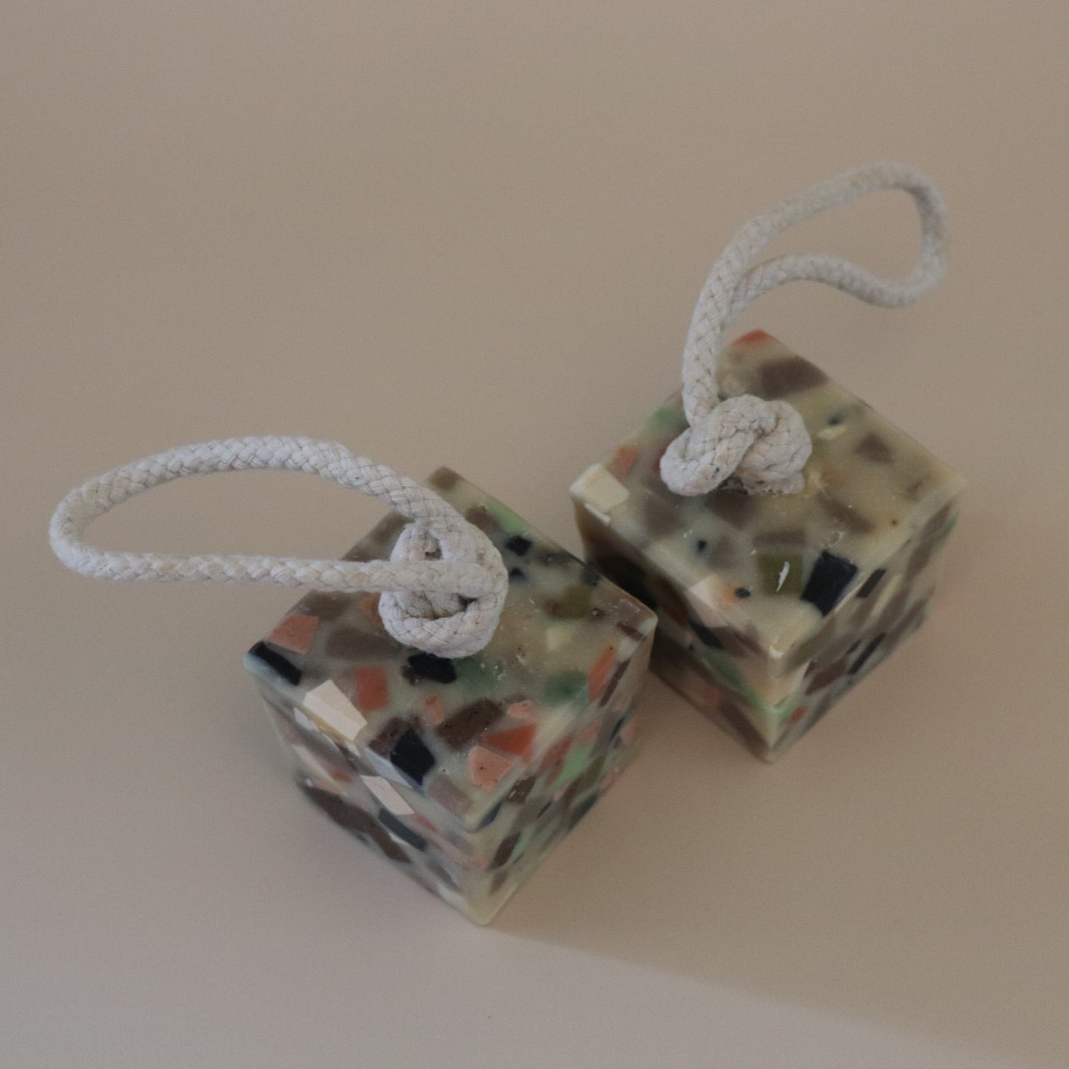 Two cube-shaped decorative terrazzo soaps with ropes attached on a beige background