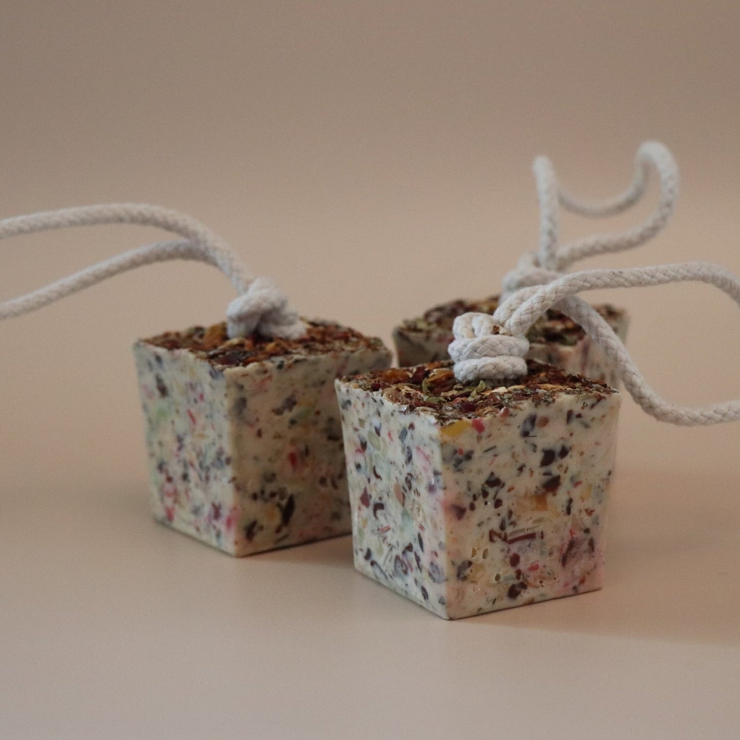 Three small cube soap bars with decorative potpourri on top and ropes attached on a beige background