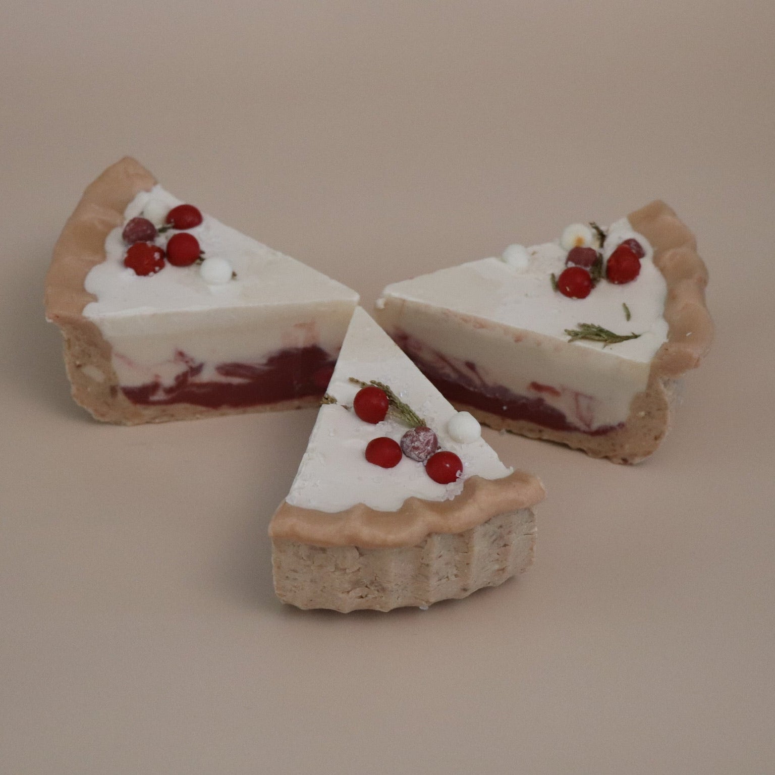 Three slices of Christmas themed pie artisan soaps with white cream and red berries on a beige background